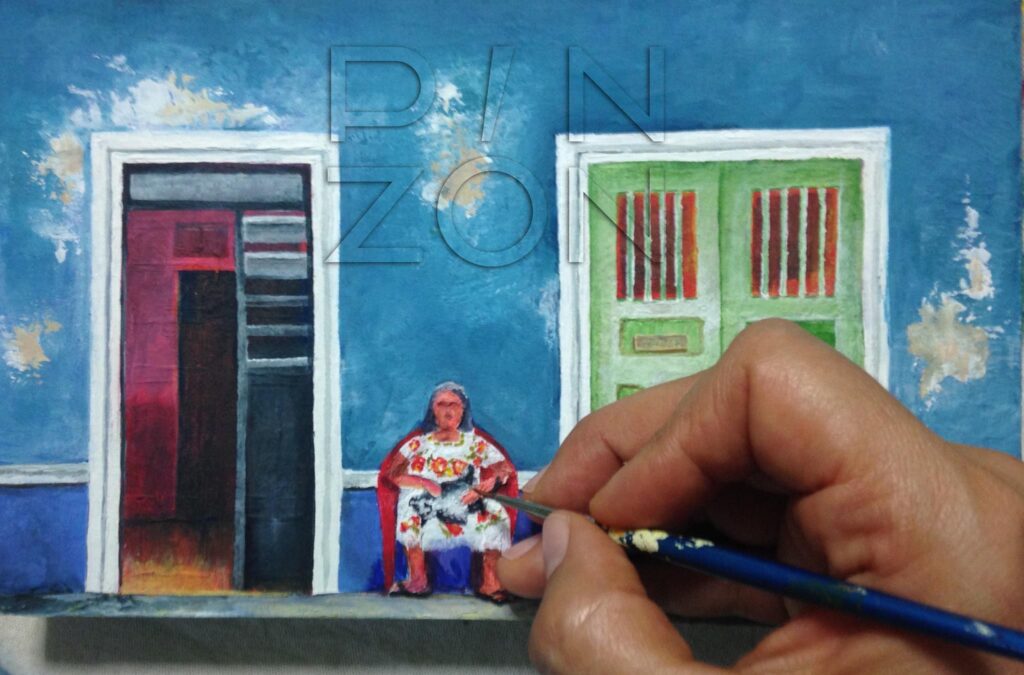 Blue traditional house in Merida City painted on a small wooden box. A woman is taking fresh air outside resting on a chair. The painter hand is shown.